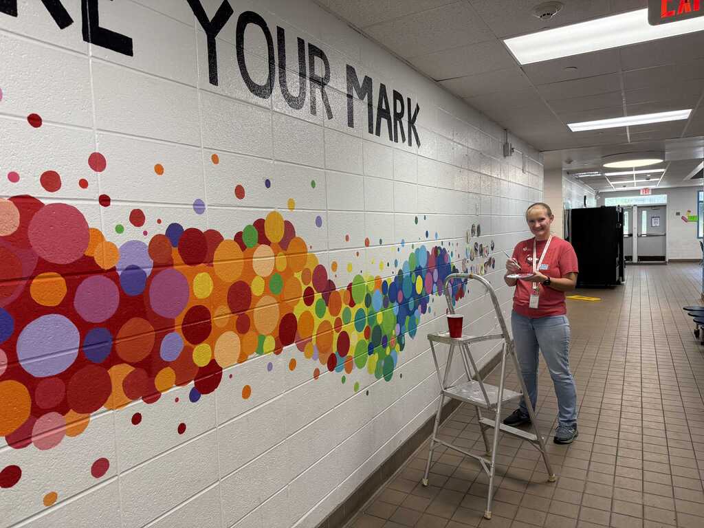 A woman stands next to a colorful mural on a hallway wall that reads "Roll Your Mark".