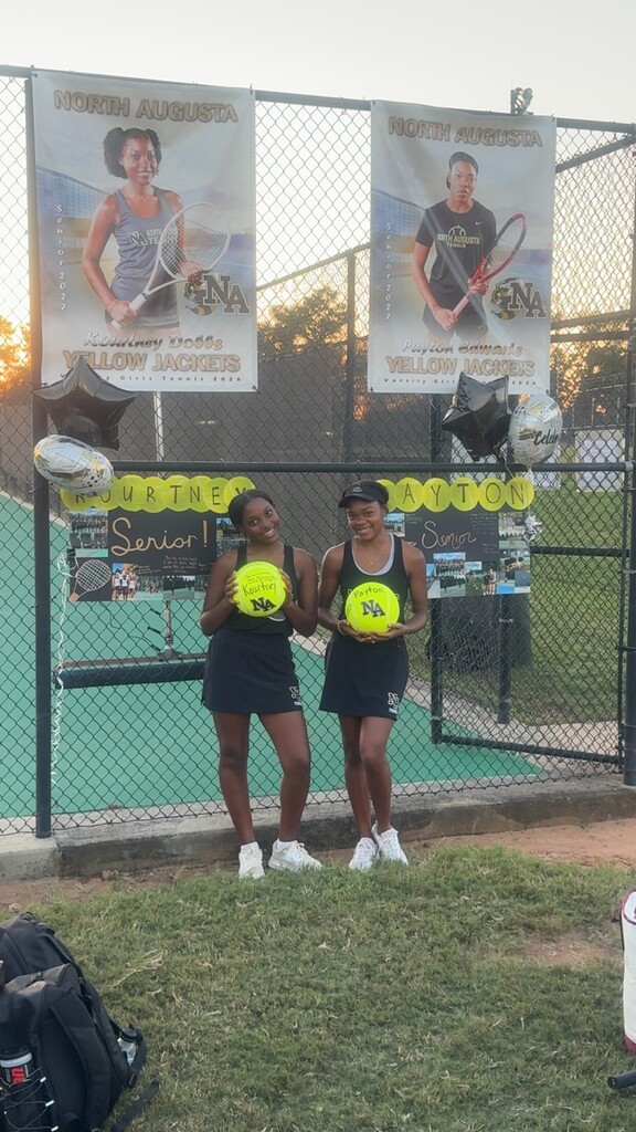 Two girls stand next to a tennis court fence holding tennis balls. Banners hang behind them. A bag sits on the grass in front.