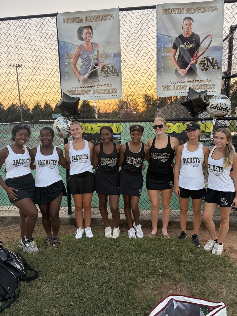 Seven women in matching uniforms stand in a row on a tennis court. Two banners hang behind them.