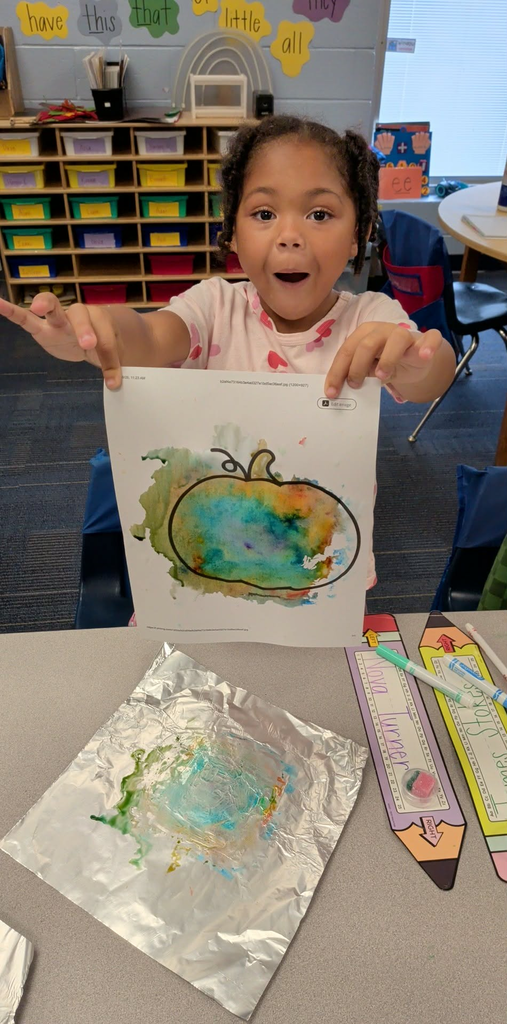 A young girl holds up a watercolor painting of a pumpkin on a white sheet of paper.