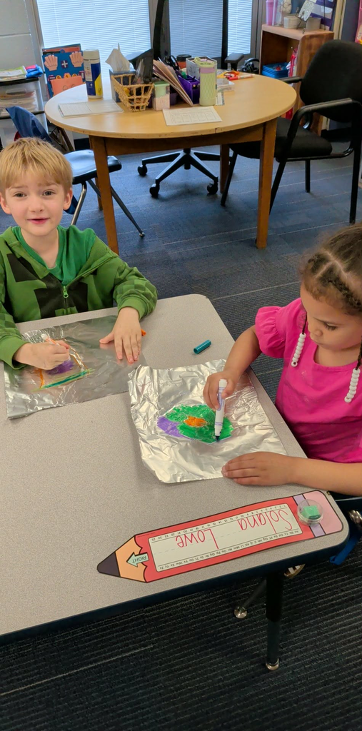Two children, a boy and a girl, seated at a table with a foil sheet. The boy is smiling and the girl holds a marker.