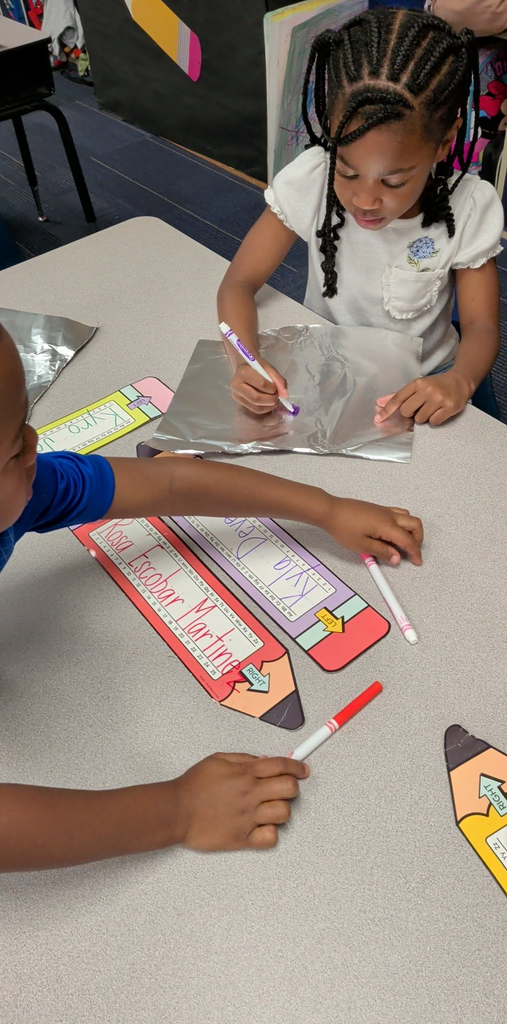 Two children, one in blue, the other in white, use markers to write on a table covered with foil and paper cutouts.