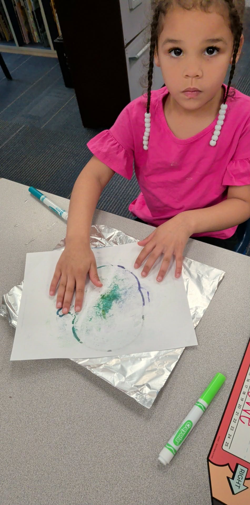 A young girl sits at a table, holding a paper with colorful paintwork. Beside her are markers and foil.