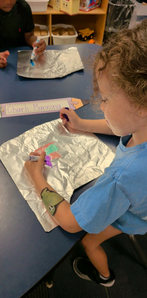 A child paints on aluminum foil with a marker at a blue table, surrounded by other students.