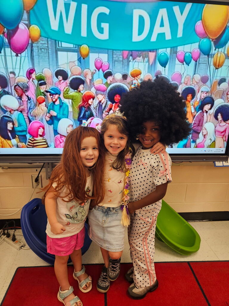 Three children pose in front of a large screen displaying "Wig Day" and various wigs. They are standing on a red carpet.