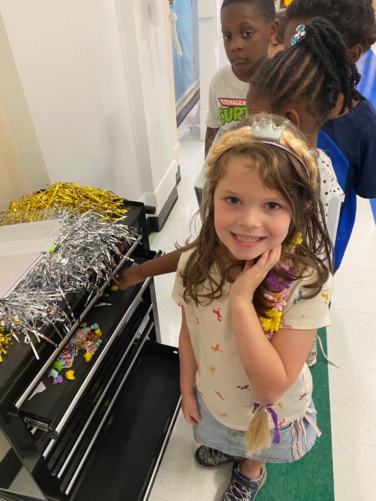 A young girl smiles next to a drawer filled with candy. Behind her are other children, one wearing a white shirt.