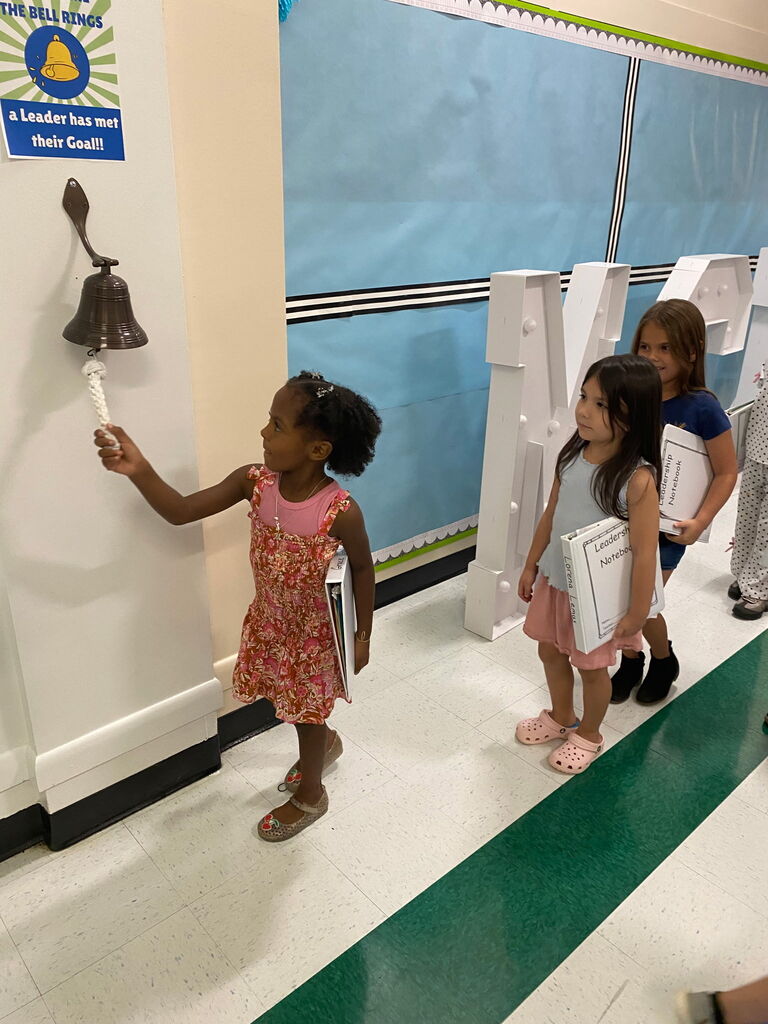 Three girls ring a bell on a wall. One girl holds a paper. The wall has a blue and white color.