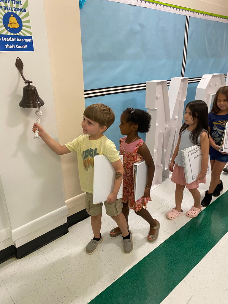 Four children, two holding books, walk in a school corridor. A boy rings a bell on a wall.