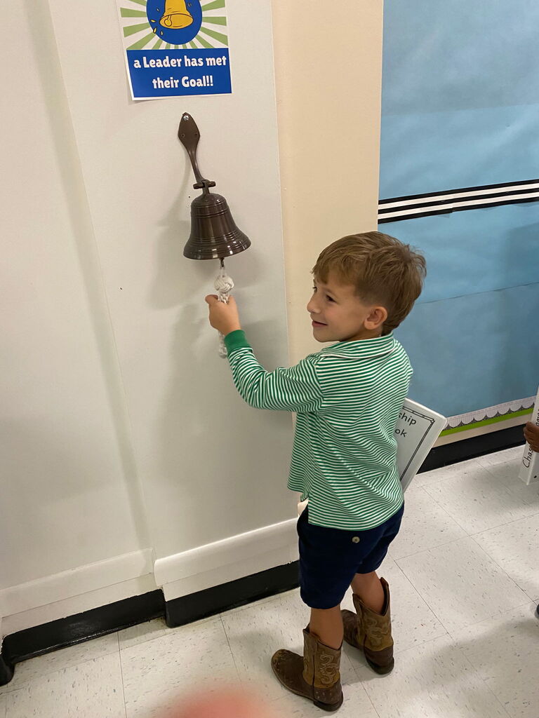 A boy with a green shirt, blue shorts, and brown boots rings a bell. He stands near a white wall.