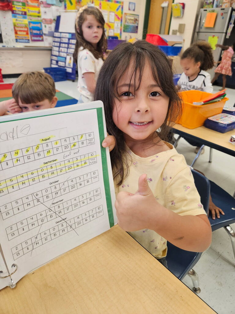 A young girl shows a book with numbers and letters, sitting at a desk in a classroom.