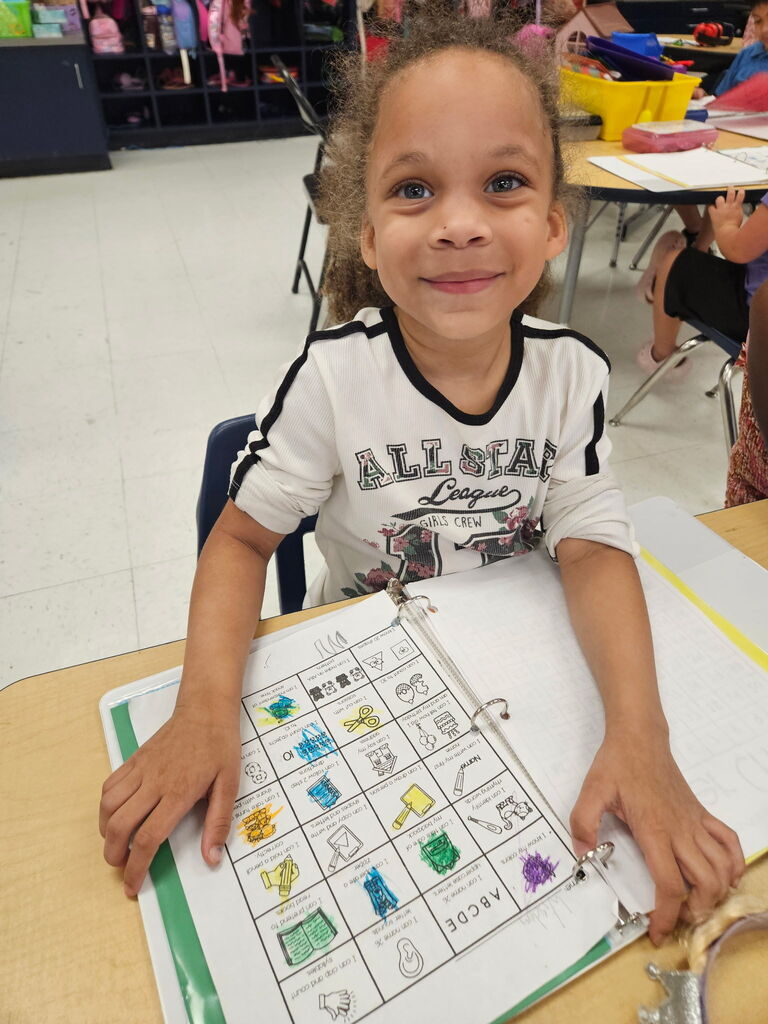 Child smiling, seated at a desk, holding an open binder with colorful pictures and numbers.