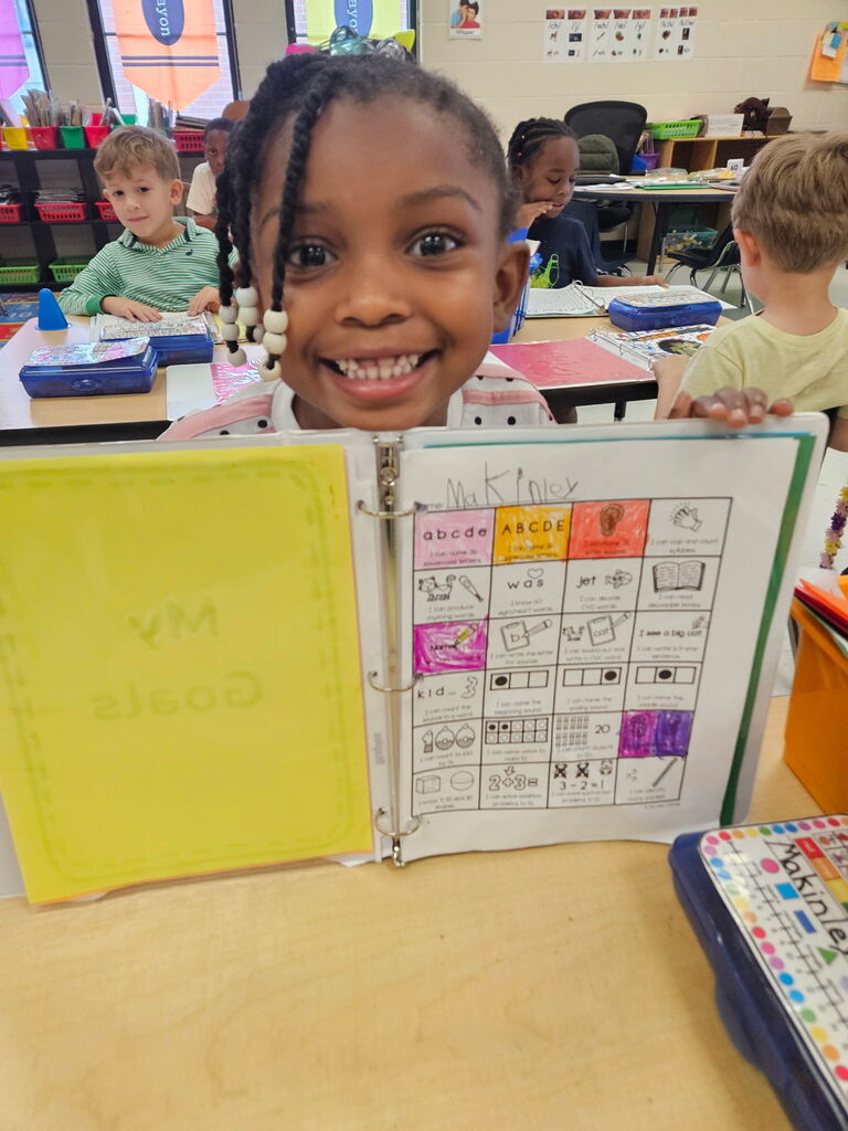 A child holds a binder with colored pages, smiling at the camera in a classroom with other children.