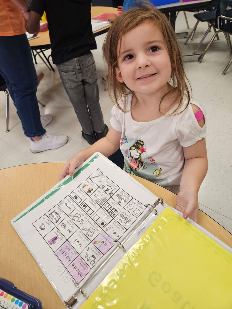 A young girl with short hair holds a binder with a yellow cover and white pages.