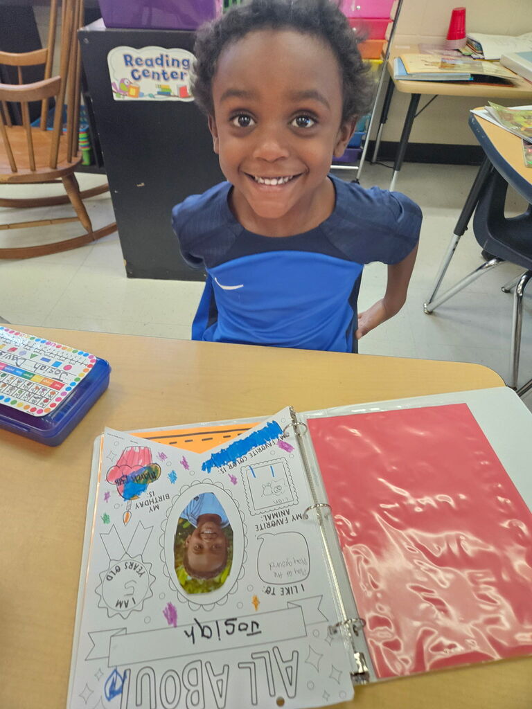 A child sitting at a desk, smiling. An open binder is in front, with a photo of the child.