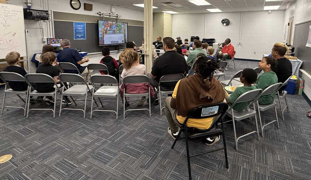 A classroom with chairs arranged in a semi-circle around a teacher at the front, with a screen at the front.