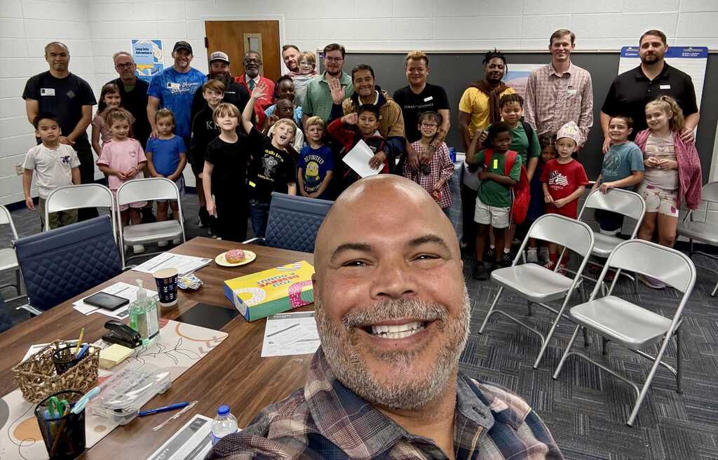 A group of people stands in a room, smiling. An adult male smiles and takes a selfie at a desk.