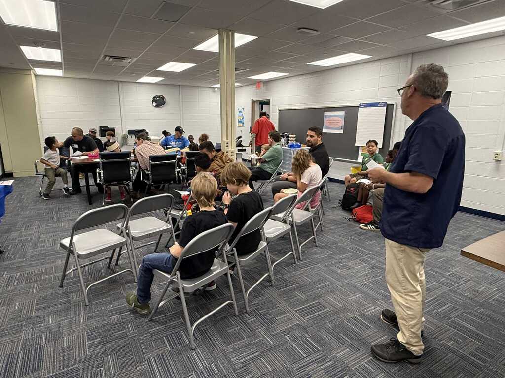 A man stands in a classroom, speaking to a group of people seated in chairs. Walls display bulletin boards and a blackboard.