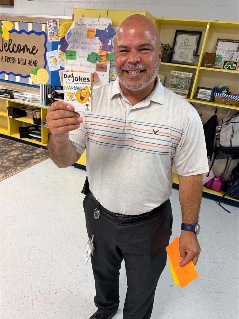 Man wearing a white shirt and black pants standing in a classroom and holding a card with a smiley face.
