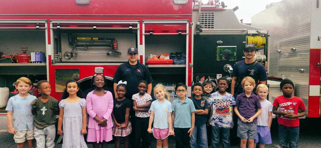 Children in light and dark clothing stand in front of a red fire truck with two firefighters.