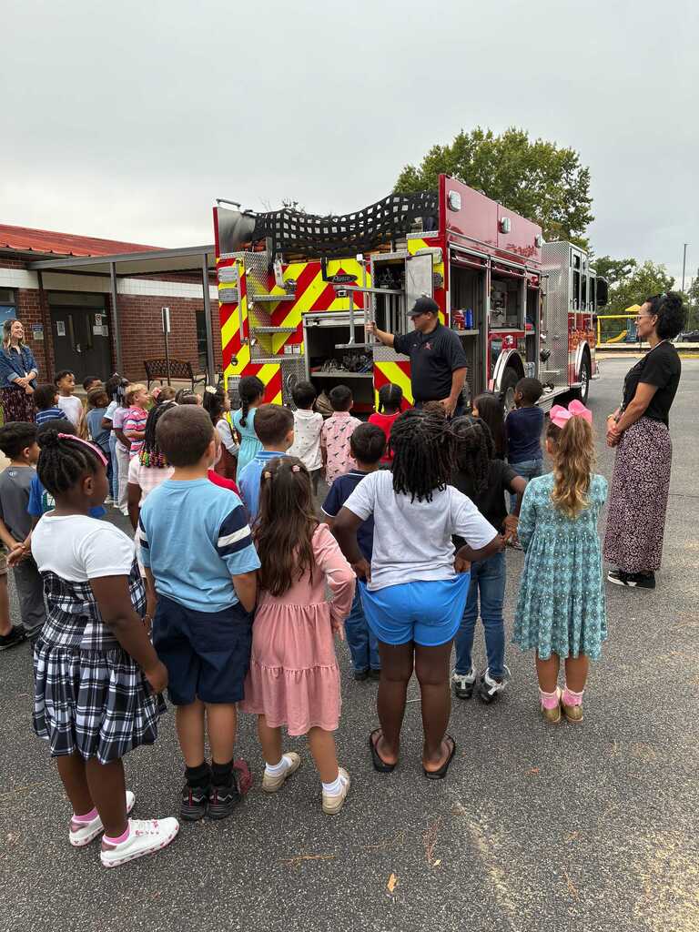Group of children stand around a fire truck; one adult guides them. A second adult stands beside the truck.