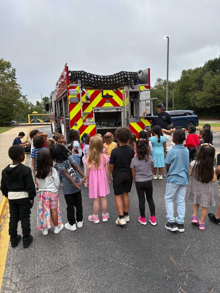 Children gathered around a fire truck with a man speaking on the ladder. Trees and a streetlight in the background.