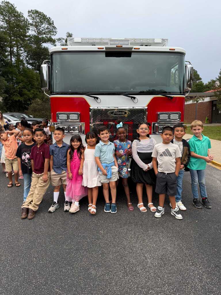 A group of children standing in front of a fire truck. Some trees are visible in the background.