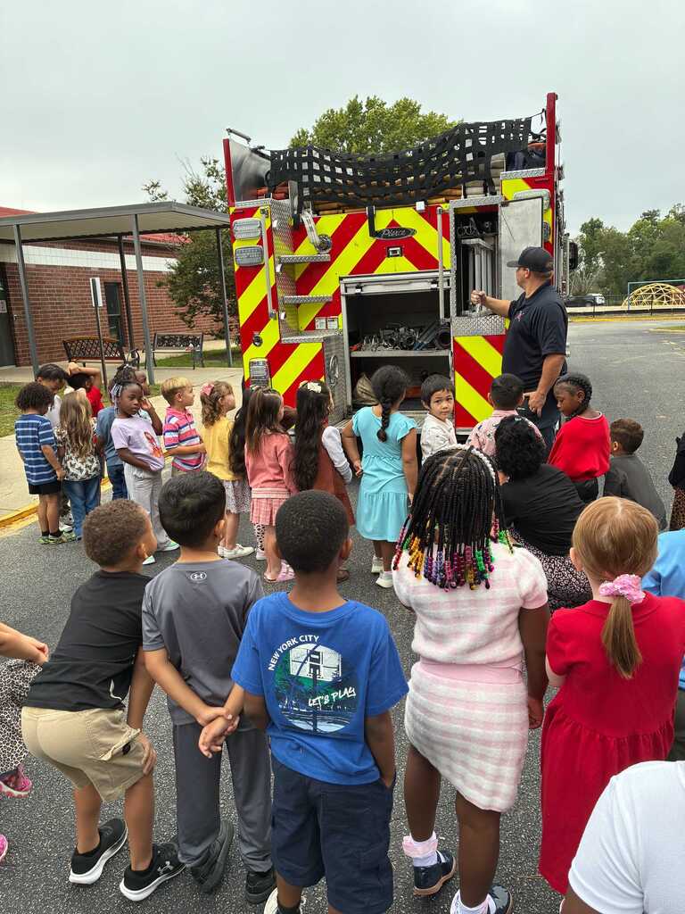 A group of children stands in a circle in a school parking lot, facing a fire truck.