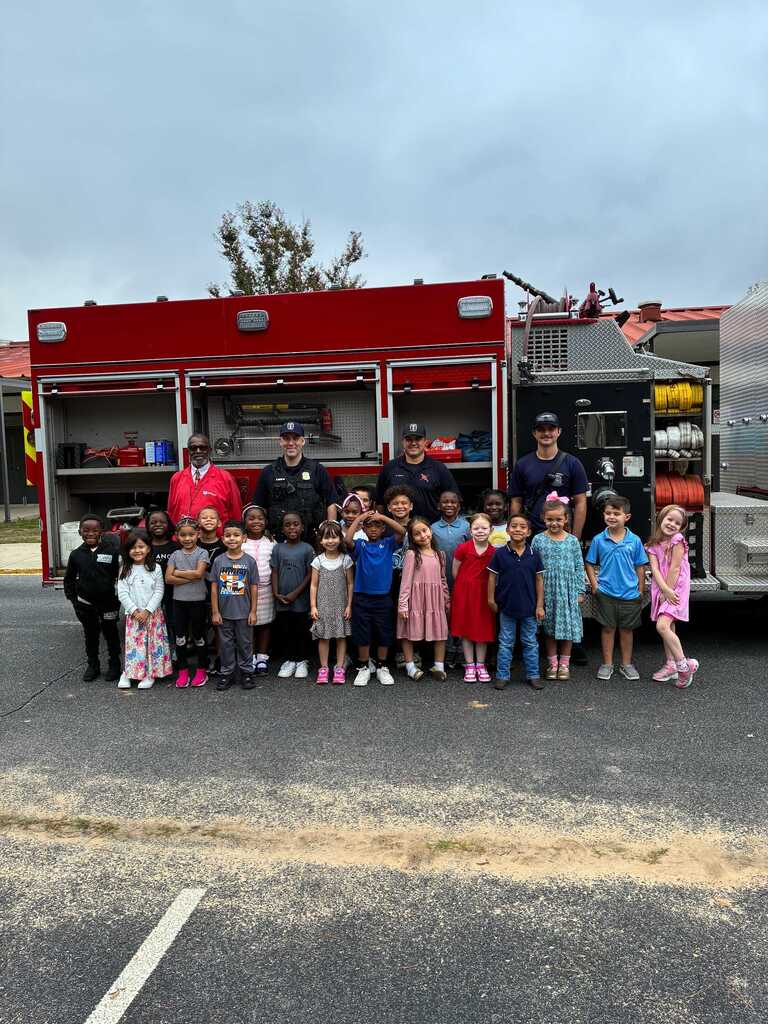A group of children, adults, and firefighters pose in front of a red firetruck.