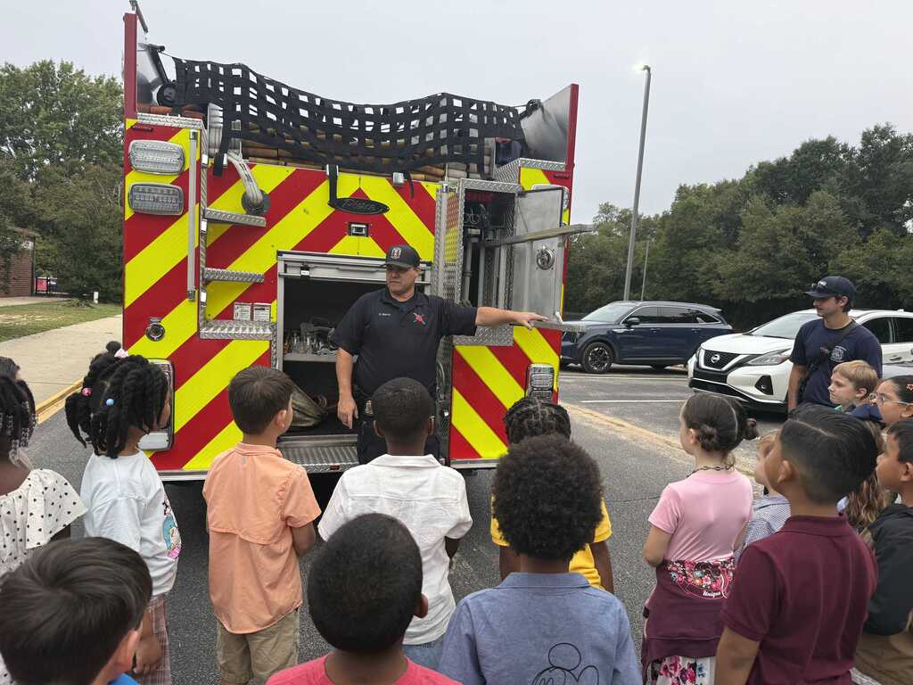 A firefighter speaks to children in a line outside a fire truck. A street is behind them with trees.