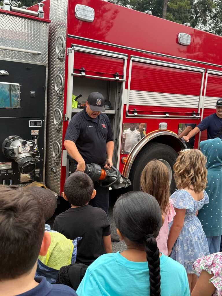 A fireman demonstrates equipment to a group of children, who attentively listen. The fire truck is parked, with the doors open.