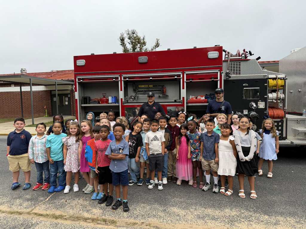 Group of children, two firefighters, and a red fire truck in a school parking lot.