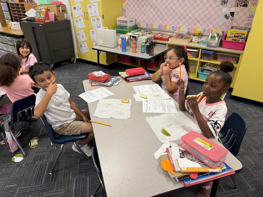 Students sit at a desk with papers, pencils, and books. Some are seated while others stand.