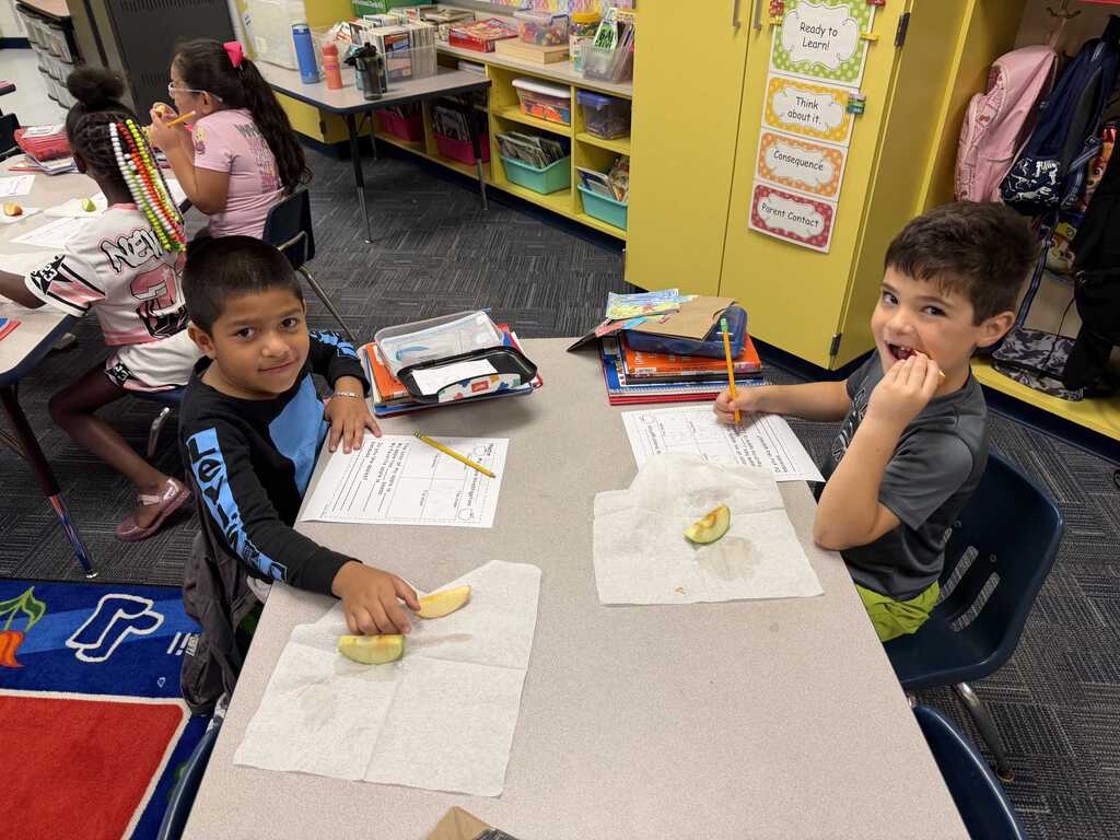 Two children sit at a desk in a classroom. They are cutting food on napkins.