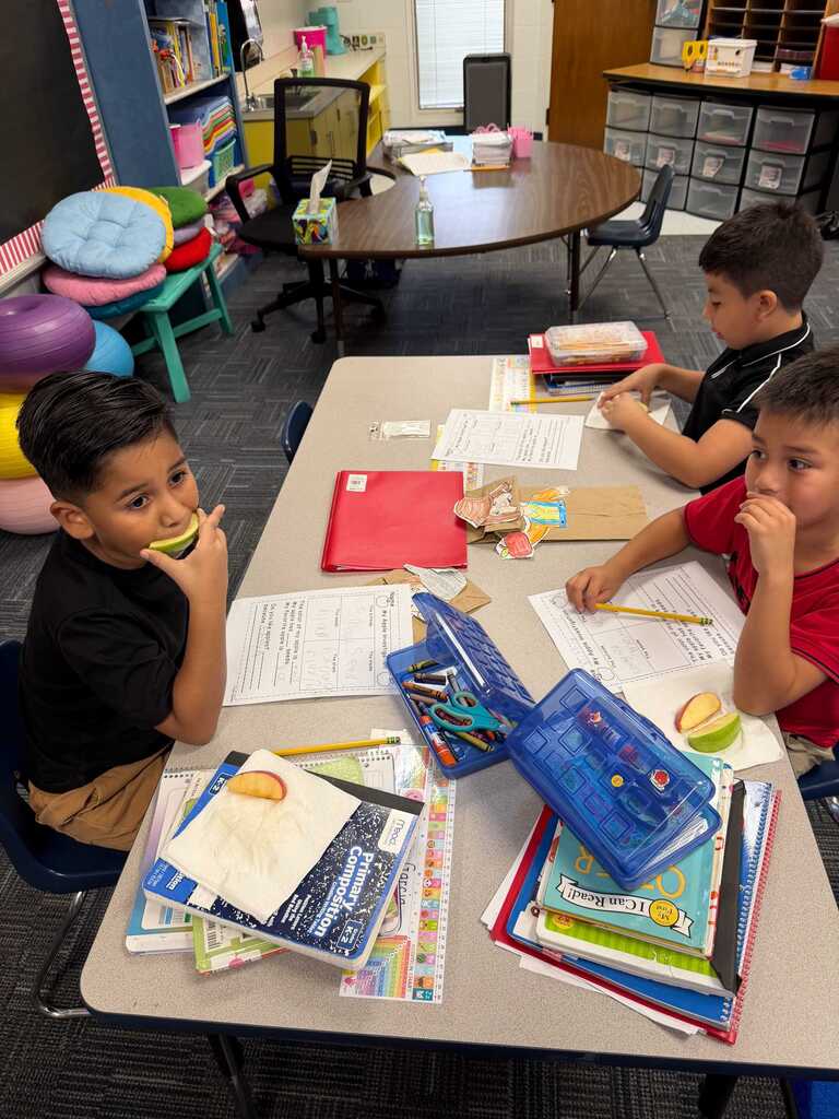Three boys at a table. One boy eats an apple, another eats a sandwich. Books and pencils are on the table.