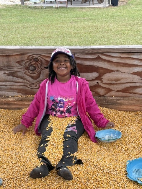 Child with pink jacket sitting in corn, holding bowl with corn kernels. Wooden wall in background.