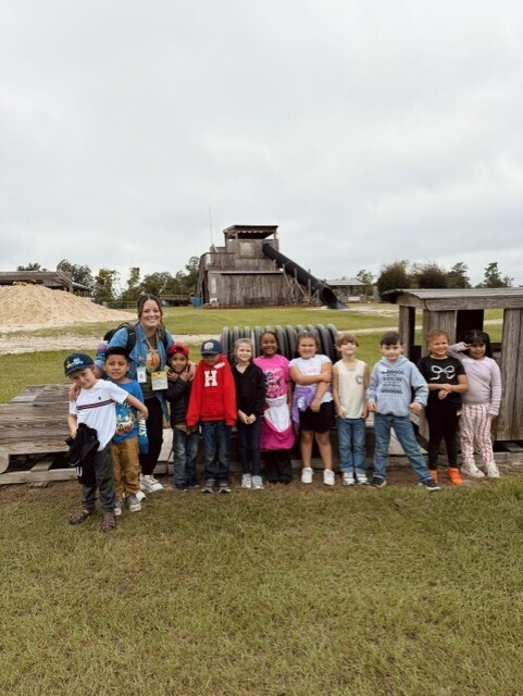 Group of children and an adult stand in front of an old wooden building, on a grassy field.