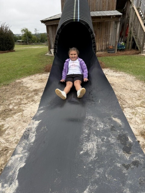 A young child sits on a black slide at a playground. Behind them is a wooden structure.