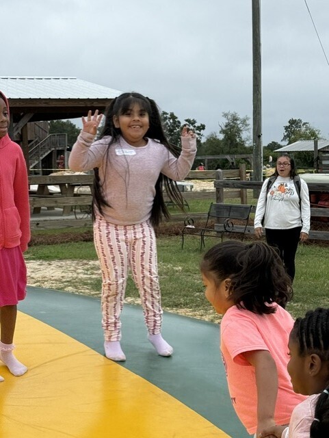 Four children play in an outdoor area. One girl jumps on a trampoline. Another girl waves.