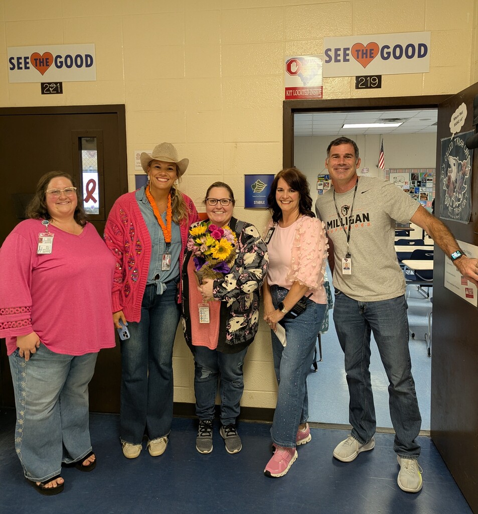 Five adults stand in a room, smiling for a photo. A woman holds flowers. Two doors are behind them.