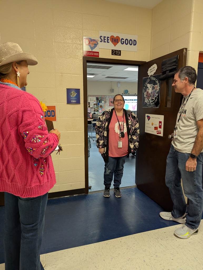 Three people in a hallway. A person wears a hat, pink sweater, and jeans. Another person wears a pink T-shirt and jeans. The last person wears a white shirt, jeans, and sneakers.