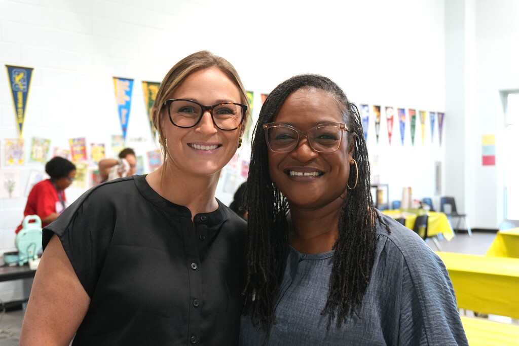 Two women with glasses smiling, standing close together. A table with a yellow tablecloth behind them.