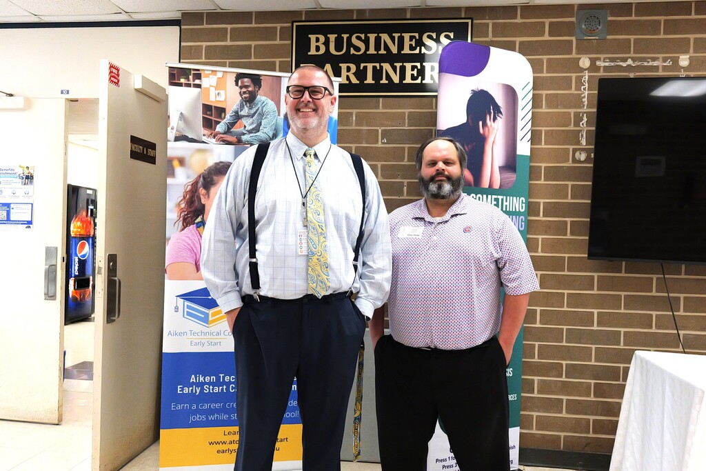Two men stand in front of a brick wall. One wears a tie, the other a polo shirt. Banners are on the wall.