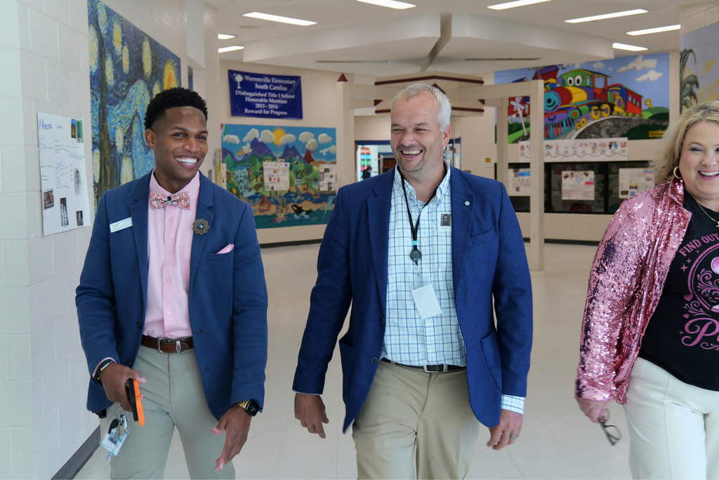 Three individuals in professional attire smile in a hallway. Brightly colored artwork adorns the walls.