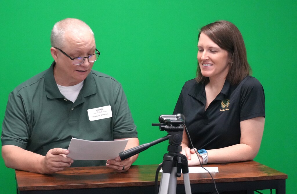 Man and woman sitting at a table, both wearing polo shirts. The man is reading from paper.