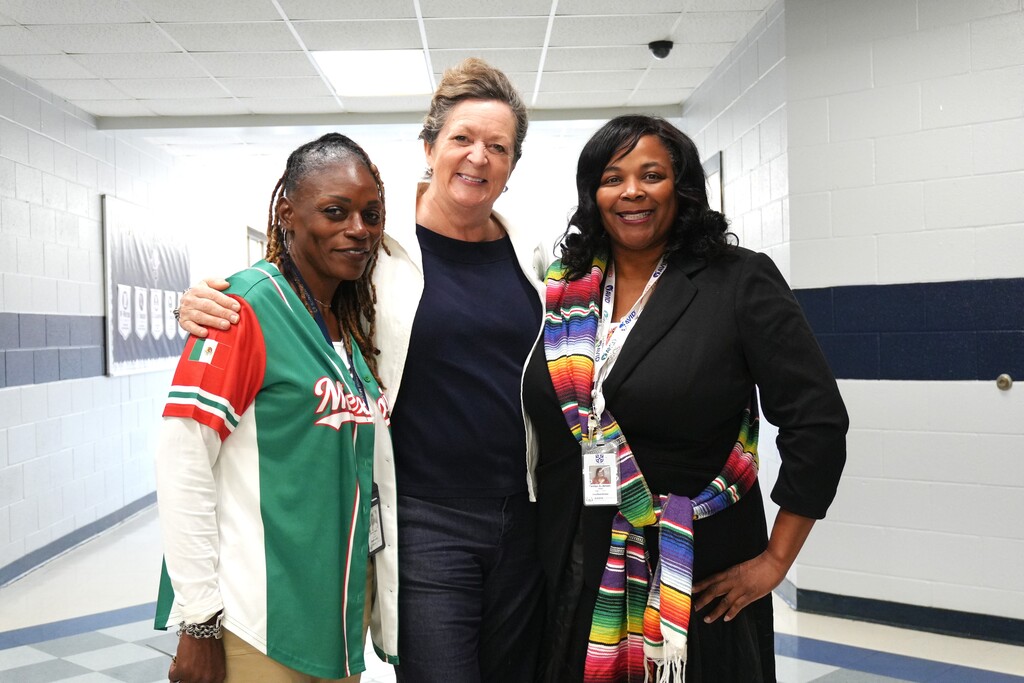 Three women stand in a corridor. The woman on the left wears a green jacket. The middle woman wears a white jacket and a name tag. The woman on the right wears a black jacket with a colorful scarf.