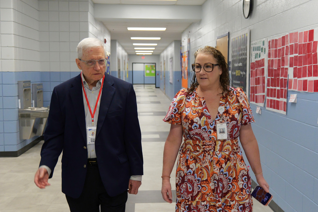 An adult man and woman walk down a brightly lit school corridor with blue walls and white tiles.