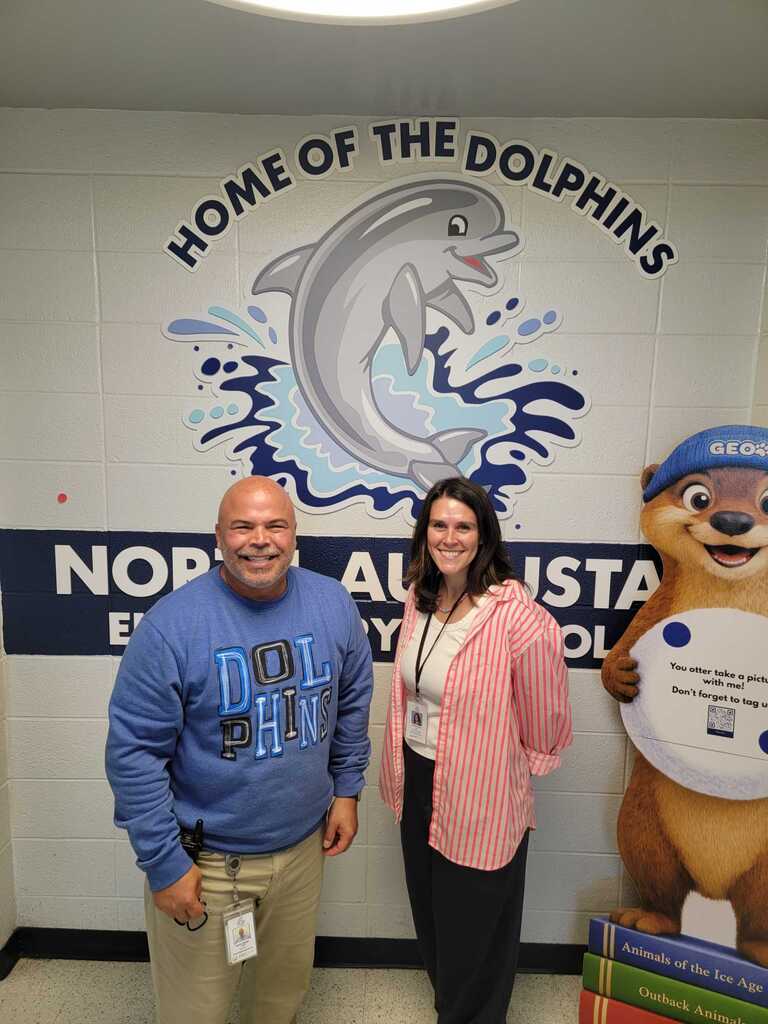 A man and woman stand in front of a dolphin mural. The man wears a blue sweatshirt. A woman wears a pink blazer. A mascot sign stands behind them.