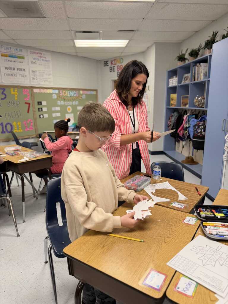 A teacher and student work at a desk in a classroom. The student is wearing glasses and has a piece of paper.