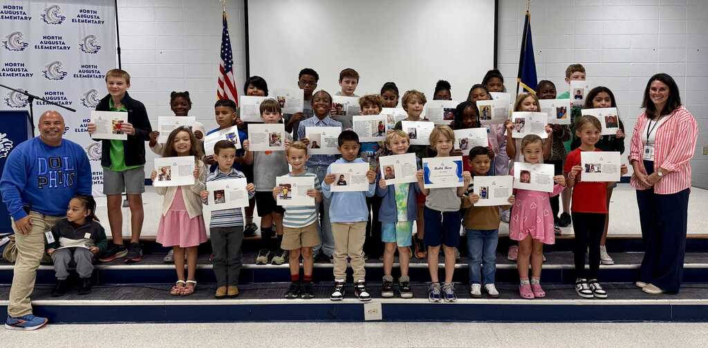 Group of children holding certificates in a room. Adults stand on a stage behind them.
