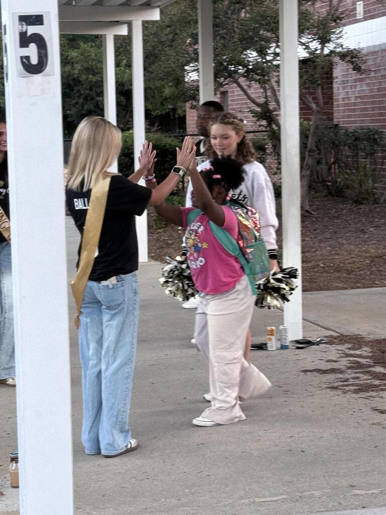 Two women and a child are high-fiving each other outside a school building. A brick wall and trees are in the background.